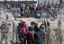 A crowd of people at a border area with law enforcement and barbed wire