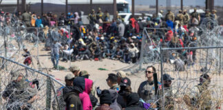 A crowd of people at a border area with law enforcement and barbed wire