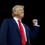 A man in a suit with a red tie raising his fist confidently during a speech