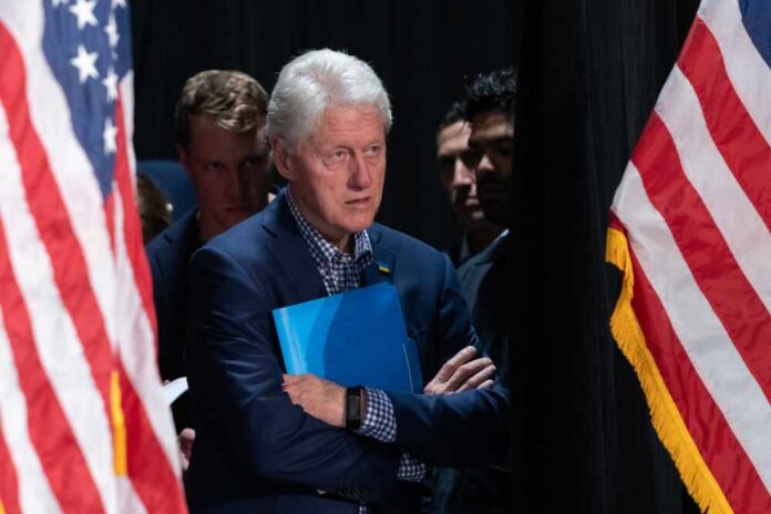 A public figure standing with arms crossed, holding a blue folder, flanked by American flags