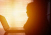 Silhouette of a person working on a laptop with sunlight streaming through blinds