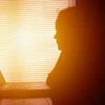Silhouette of a person working on a laptop with sunlight streaming through blinds