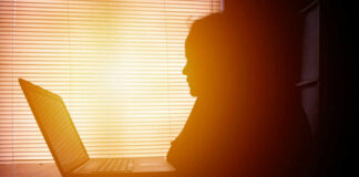 Silhouette of a person working on a laptop with sunlight streaming through blinds