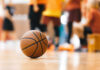 A basketball resting on the gym floor with players blurred in the background