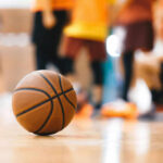 A basketball resting on the gym floor with players blurred in the background