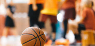 A basketball resting on the gym floor with players blurred in the background