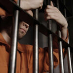 A male prisoner in an orange jumpsuit holding onto the bars of a jail cell