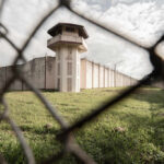 View of a prison tower surrounded by a security fence and grassy area under a cloudy sky