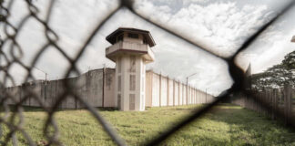 View of a prison tower surrounded by a security fence and grassy area under a cloudy sky