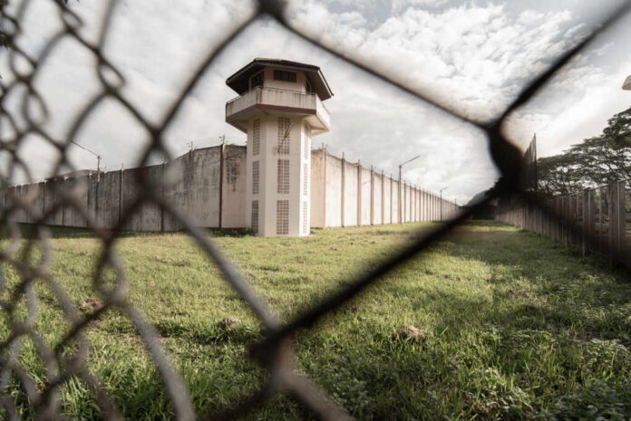 View of a prison tower surrounded by a security fence and grassy area under a cloudy sky