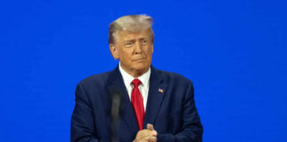Man in a suit clapping hands on a stage with a blue background