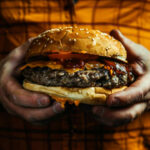 A person holding a large cheeseburger with lettuce, tomato, and condiments