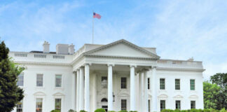 The White House with an American flag flying above, surrounded by greenery
