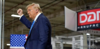A man in a suit raises his fist while standing in a manufacturing facility with an American flag in the background