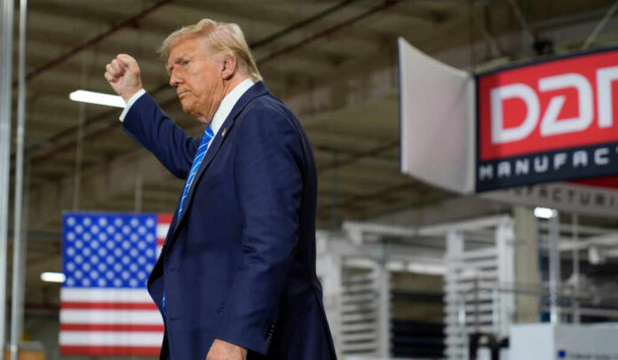 A man in a suit raises his fist while standing in a manufacturing facility with an American flag in the background