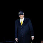 A man in a black coat and yellow tie wearing a campaign hat stands against a dark background