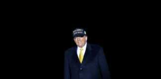 A man in a black coat and yellow tie wearing a campaign hat stands against a dark background