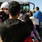 A parent holding a child in line near a bus with other individuals waiting