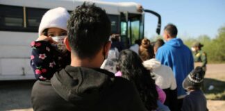 A parent holding a child in line near a bus with other individuals waiting