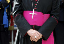 A bishop in traditional attire with a cross necklace and a pink sash, hands clasped in front