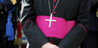 A bishop in traditional attire with a cross necklace and a pink sash, hands clasped in front