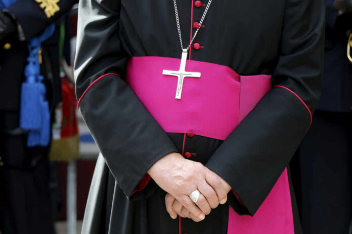 A bishop in traditional attire with a cross necklace and a pink sash, hands clasped in front