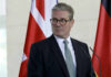 80 MPs Demand UK Prime Minister’s Resignation A man in formal attire standing at a podium with flags in the background