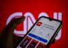 A hand holding a smartphone displaying the CNN news app against a red background