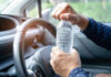 A person opening a water bottle while holding the steering wheel of a car
