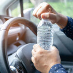 A person opening a water bottle while holding the steering wheel of a car