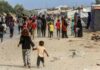 A crowded scene in a refugee camp with children and adults walking along a dusty path