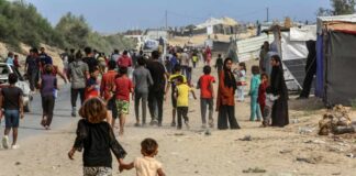A crowded scene in a refugee camp with children and adults walking along a dusty path