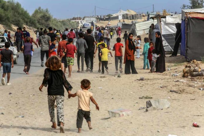 A crowded scene in a refugee camp with children and adults walking along a dusty path