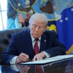 A person in a suit signing documents at a desk in the Oval Office