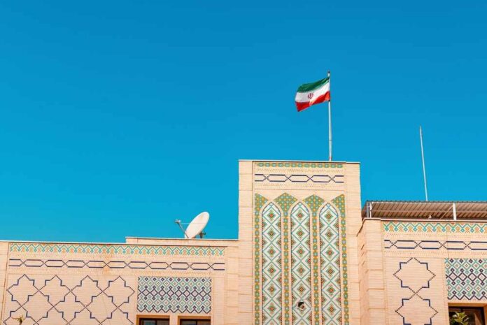 Iran,Flag,Waving,On,A,Pole,Above,An,Architectural,Facade Building with intricate patterns and the Iranian flag against a clear blue sky