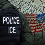 A police officer in a tactical vest stands near a barbed wire fence with an American flag in the background