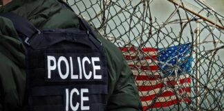 A police officer in a tactical vest stands near a barbed wire fence with an American flag in the background