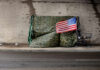 Audit Stunner: Camps Bounce Back A makeshift shelter draped with an American flag next to a shopping cart on a city street