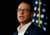 Results-First Governor Sparks Democratic Brawl A man in a suit speaking at a podium with an American flag in the background