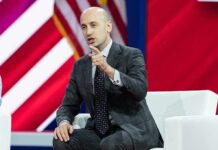 Budget Brawl Erupts Over Migrant Costs Man in a gray suit speaking on stage