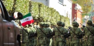 Soldiers in green camouflage march carrying rifles and a Mexican flag