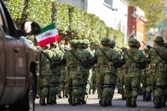 Soldiers in green camouflage march carrying rifles and a Mexican flag