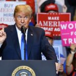 Donald Trump speaking at a rally podium with crowd holding signs