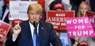 Donald Trump speaking at a rally podium with crowd holding signs