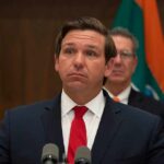 Ron DeSantis at a press conference podium in a suit and red tie