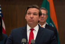 Ron DeSantis at a press conference podium in a suit and red tie