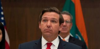 Ron DeSantis at a press conference podium in a suit and red tie